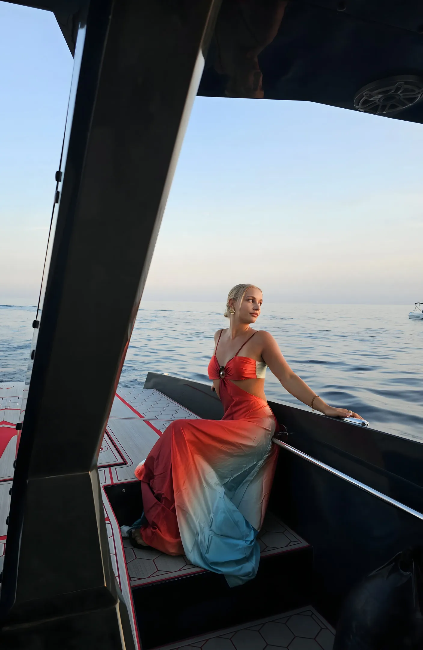 Een vrouw in een lange kleurverloopjurk zit in de schemering op de rand van de boot, achter haar de kalme open zee
